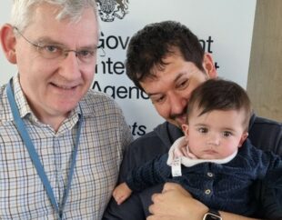 A photo of Steve holding his baby Iris and standing next to his colleague Simon on a visit to the GIAA office in London. The GIAA crest is seen behind them.